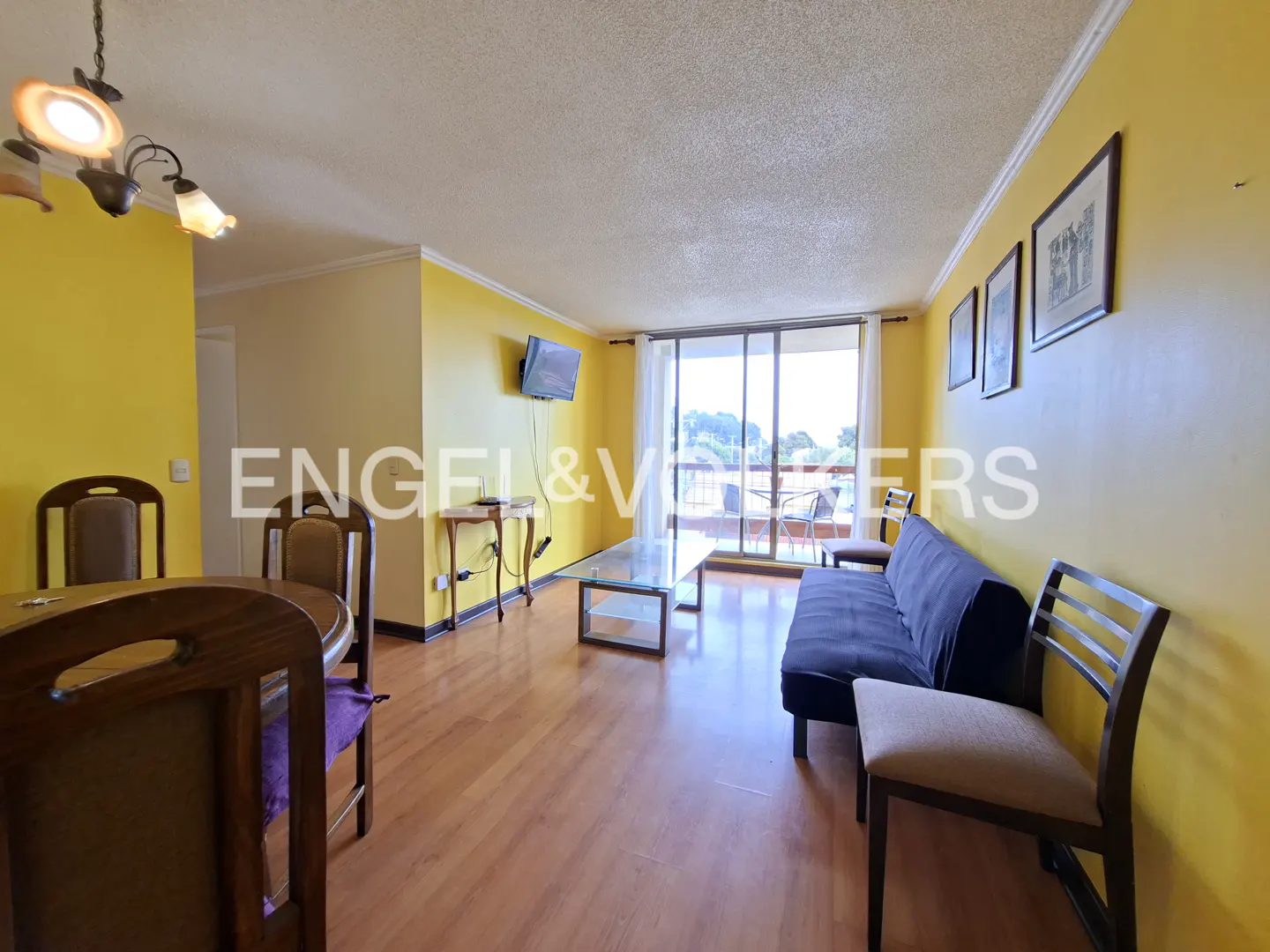 Bright living room with yellow walls, wood floors, and a glass table. A blue sofa faces a balcony with a view. Dining table and chairs in the foreground.