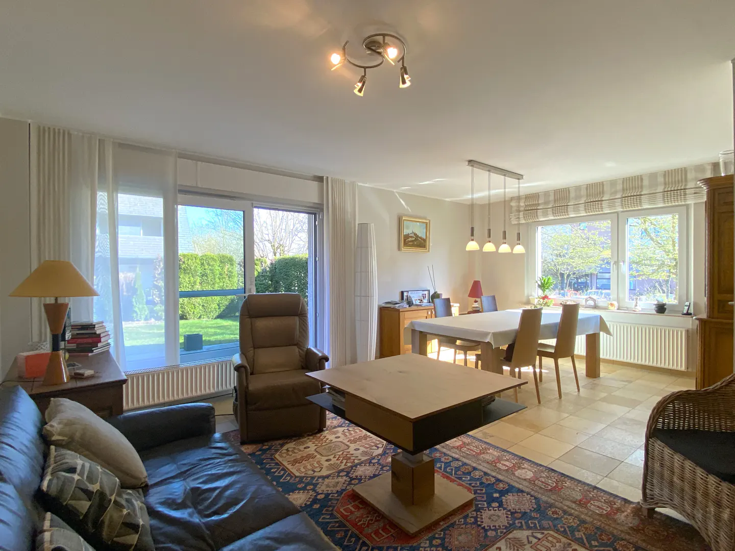 Bright living and dining room with a leather sofa, brown recliner, and a dining table set for four. A patterned rug adds warmth.
