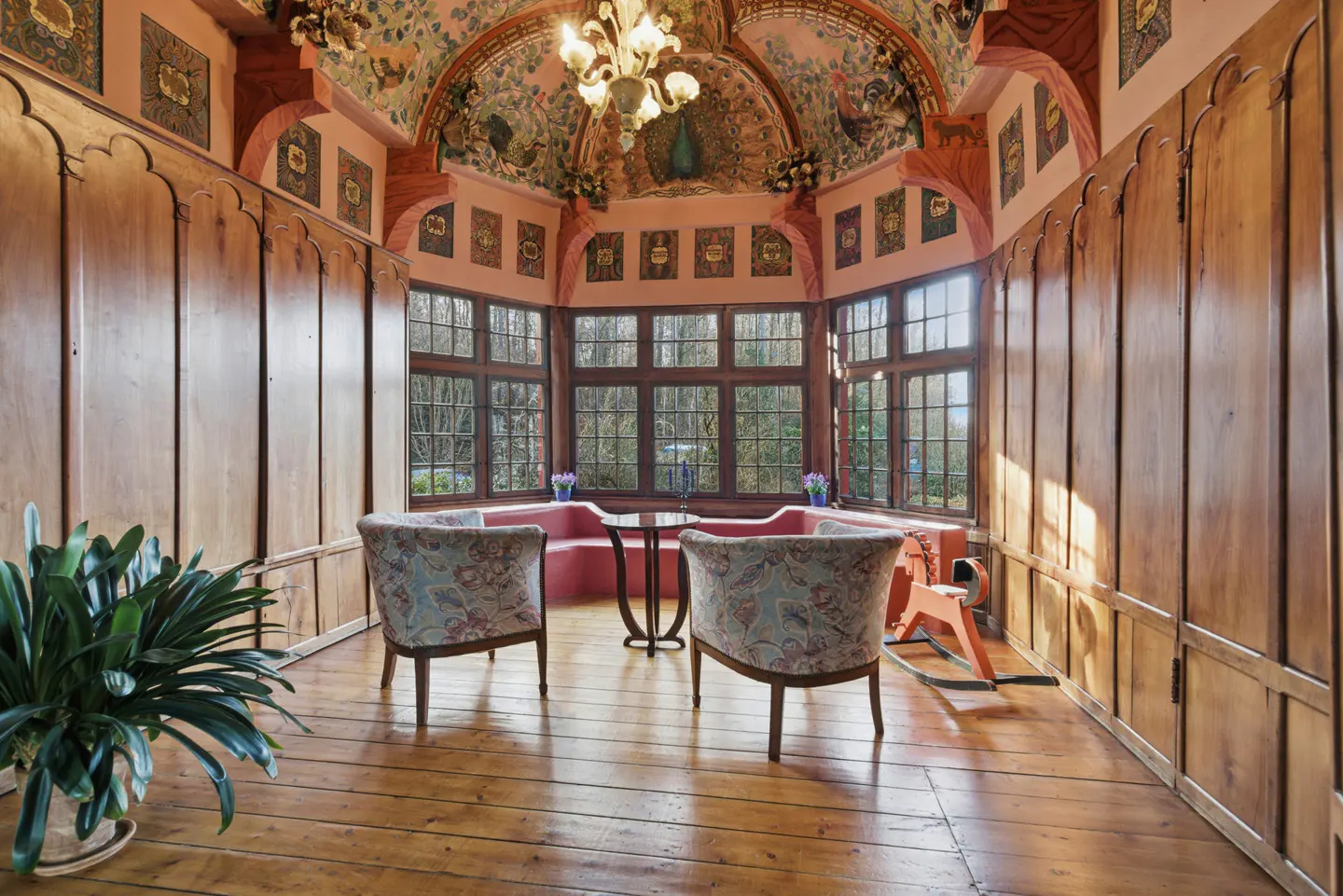 Ornate room with wood paneling, floral ceiling art, and bay window. Two floral armchairs face a small table and built-in bench.