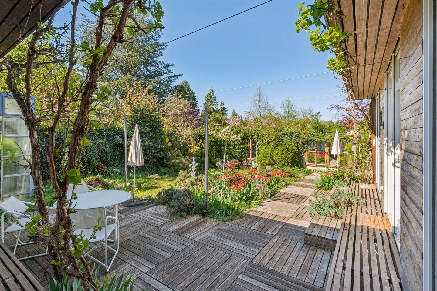 Wooden deck patio with white chairs, a small table, and a lush garden with blooming red flowers under a clear blue sky.