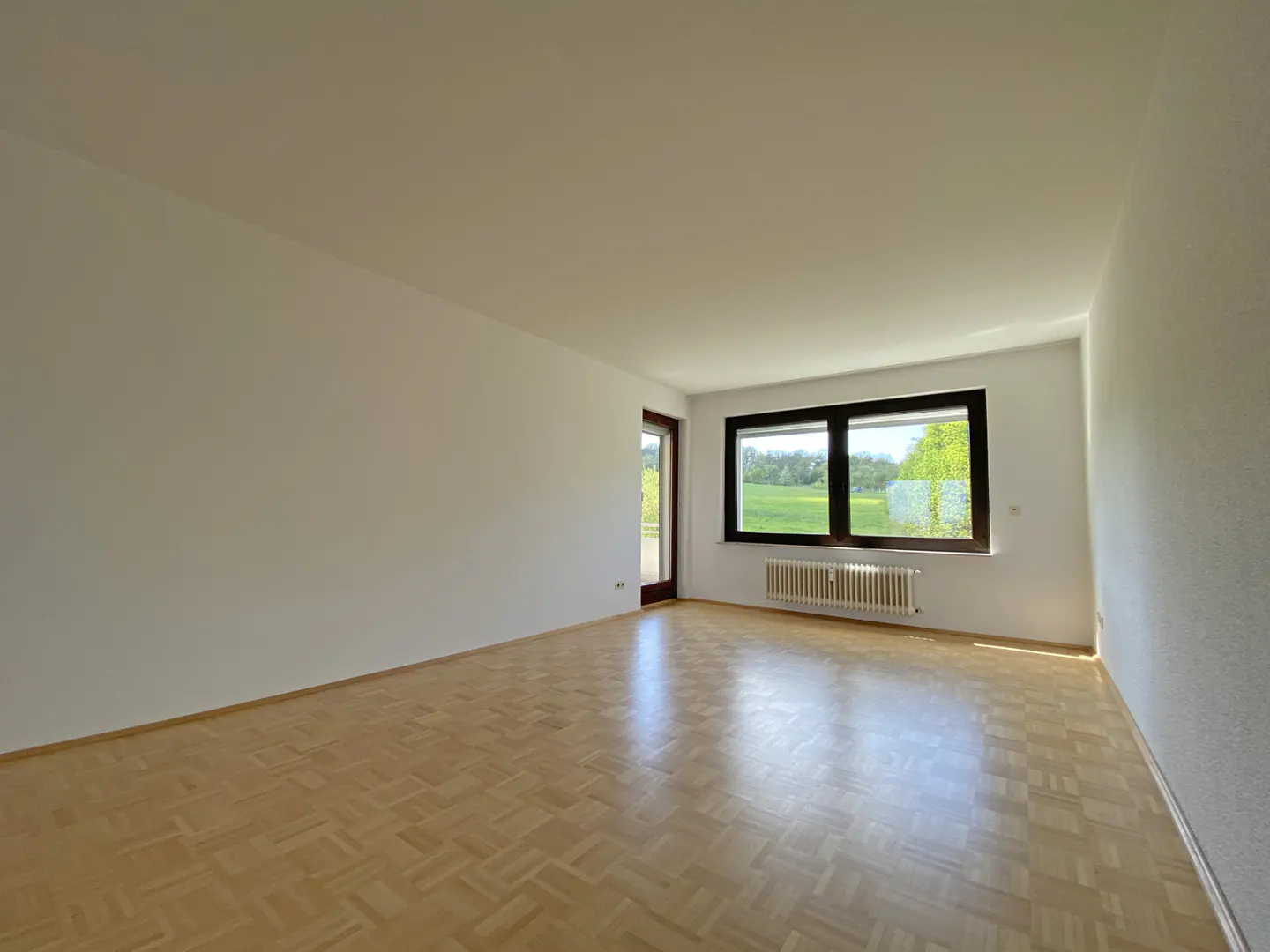 Bright, empty room with parquet floors, white walls, and a large window with a view of a green field. A radiator sits beneath the window.
