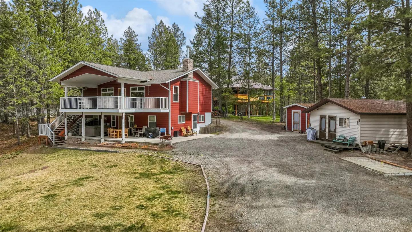 Two-story red house with a white balcony and a gravel driveway surrounded by tall green trees.
