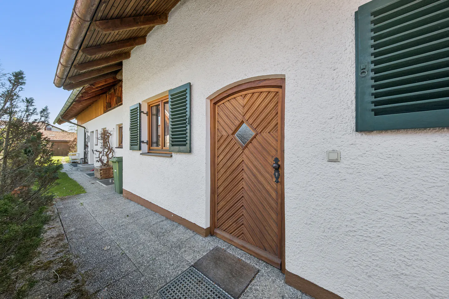 Exterior view of a white house with a wooden door and green shutters. A stone walkway leads to the entrance.