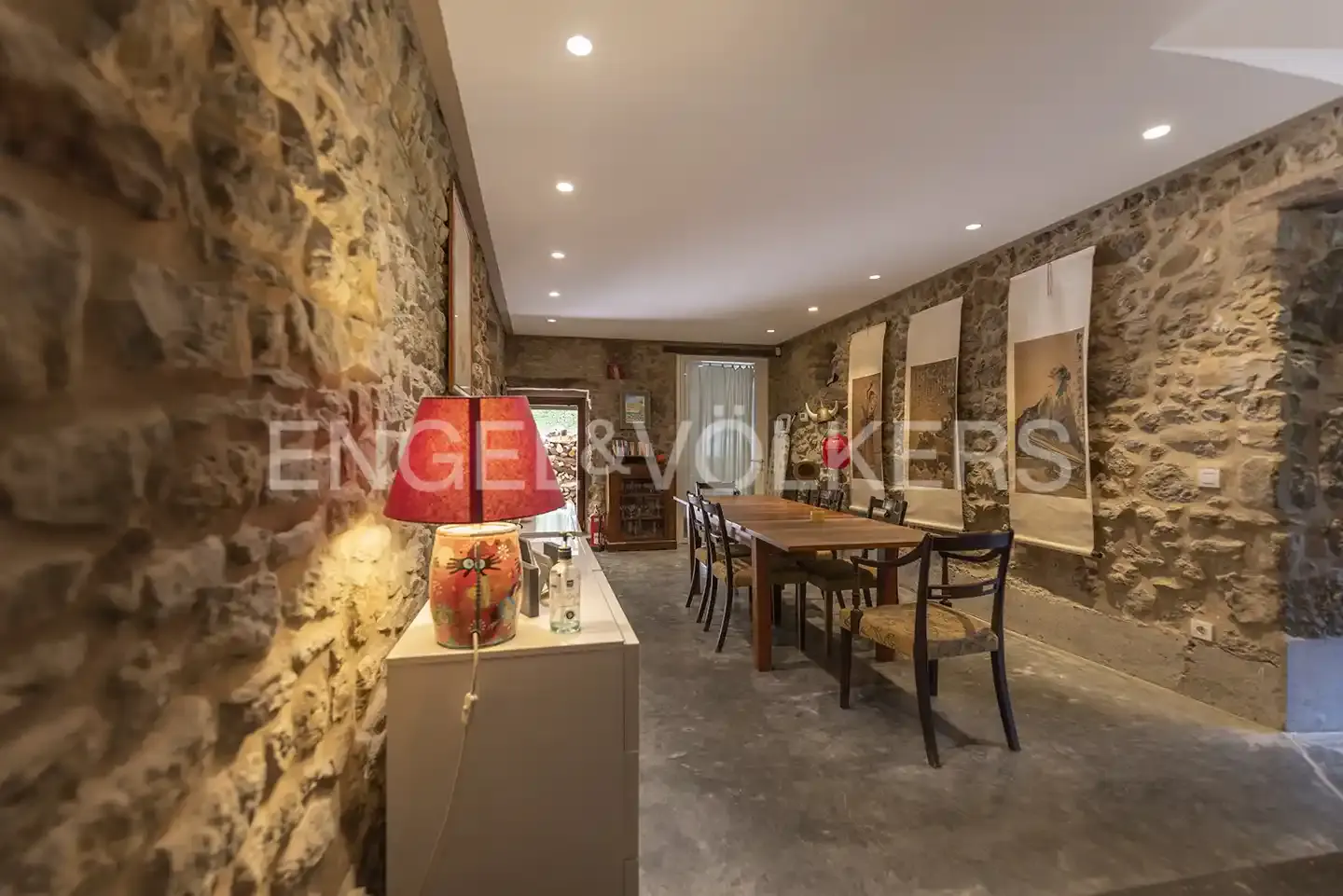 Stone-walled dining room with a long wooden table and chairs. A lamp sits on a white cabinet. Art hangs on the walls.