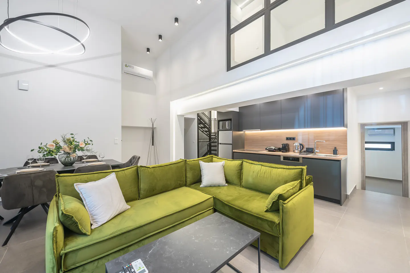 Bright, modern living space with a green velvet sectional sofa, dining table, and gray kitchen cabinets.