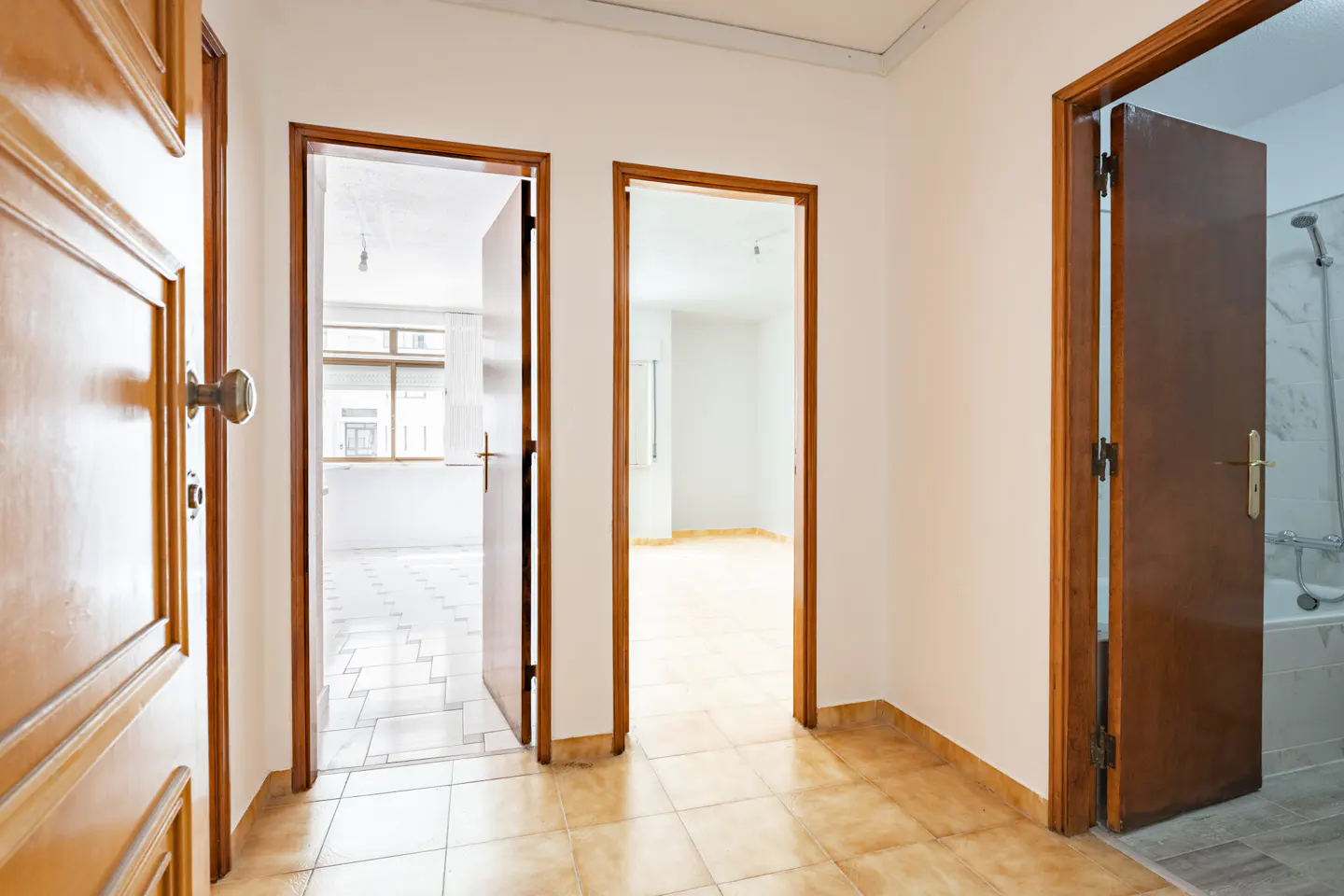 Hallway with three open wood doors. The floor is tiled in tan. The walls are white. The first door is ajar, the second and third are fully open.