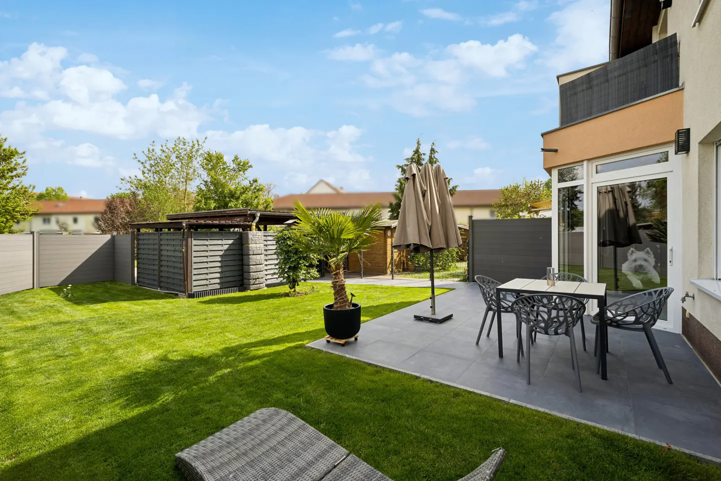 A backyard with green grass, a patio with a table and chairs, and a closed umbrella.
