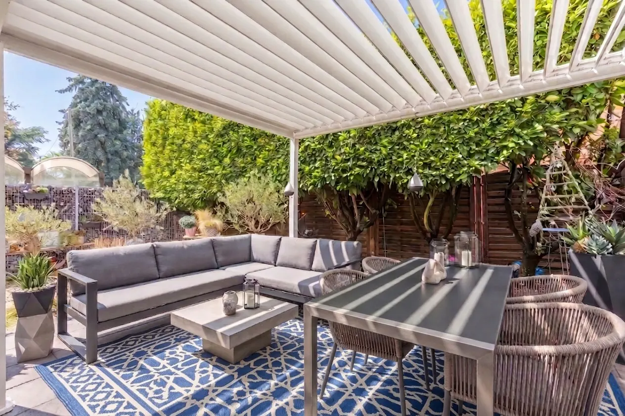 Outdoor patio with gray sectional sofa, coffee table, dining table, and chairs under a white pergola, with a blue patterned rug.