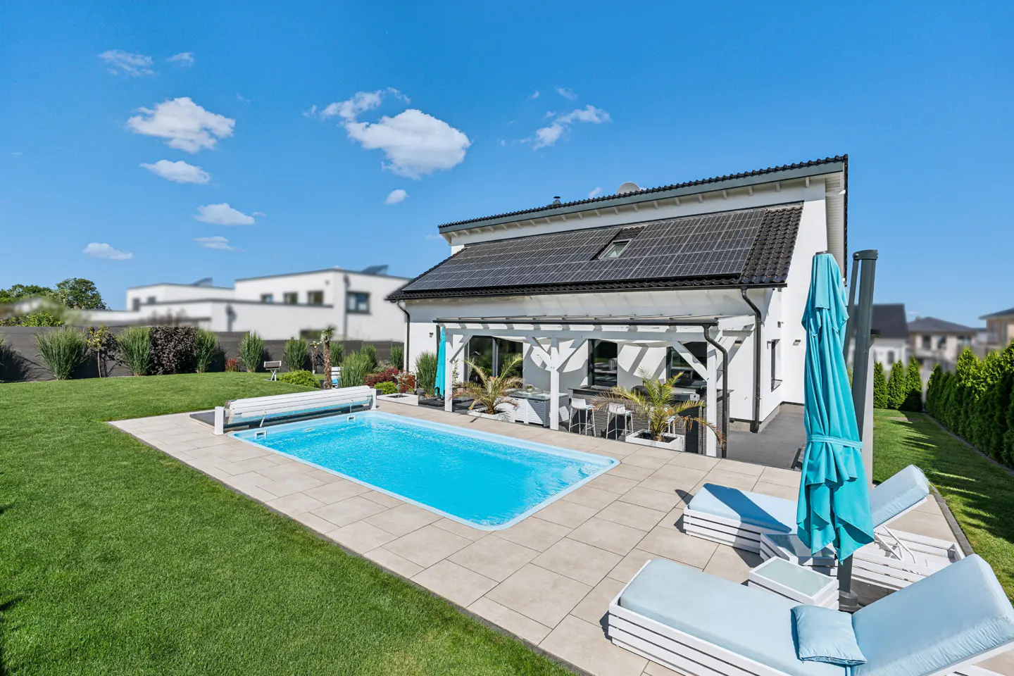 Backyard view of a modern white house with a pool, patio, and solar panels under a blue sky.