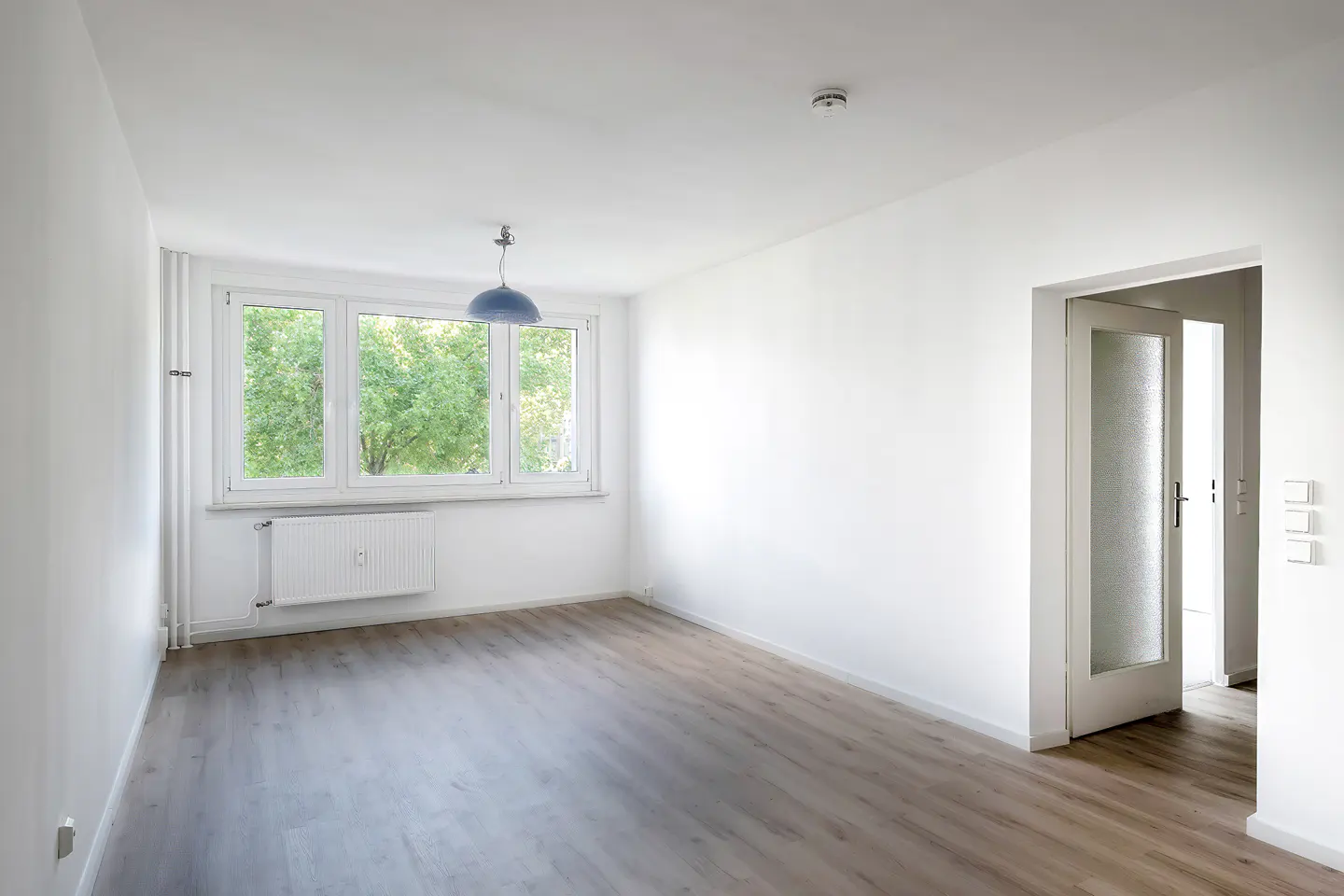 Empty room with white walls, light wood-look flooring, a window with a view of trees, and a doorway leading to a hallway.
