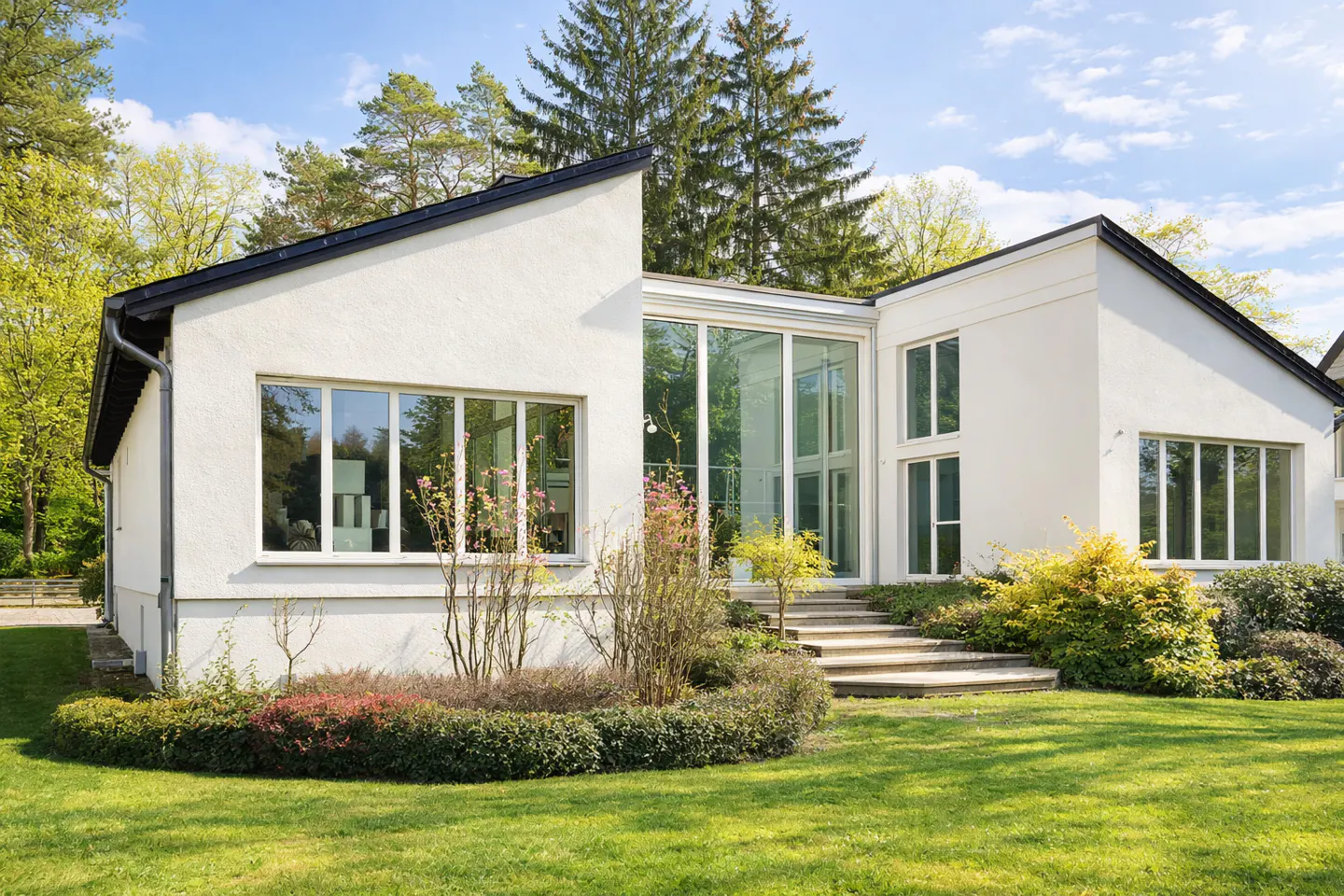 Modern white house with black roof, large windows, and glass entrance. Green lawn and trees surround the property.