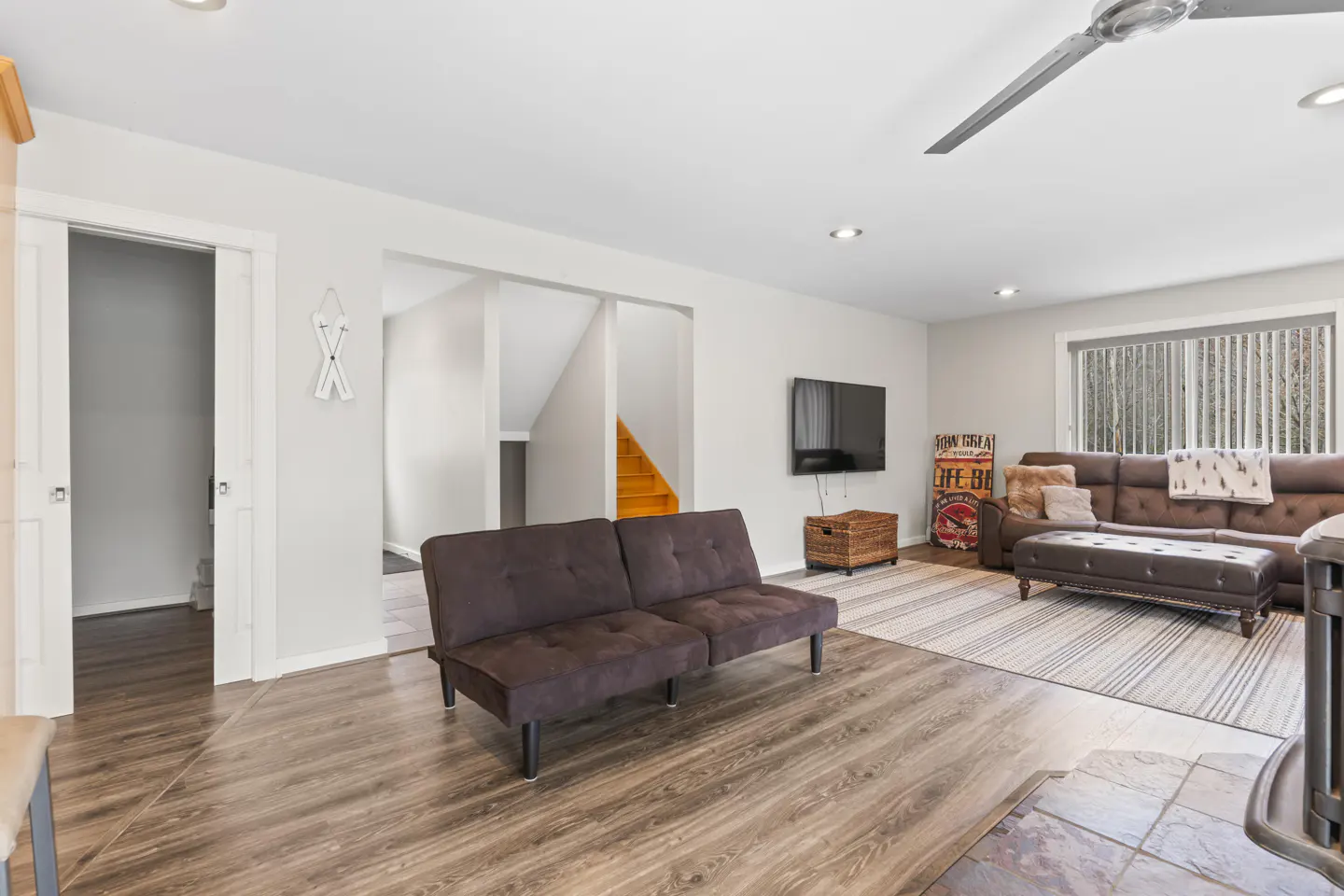 Living room with brown futon, sofa, and wood floors. A TV hangs on the wall, and stairs lead to another level.