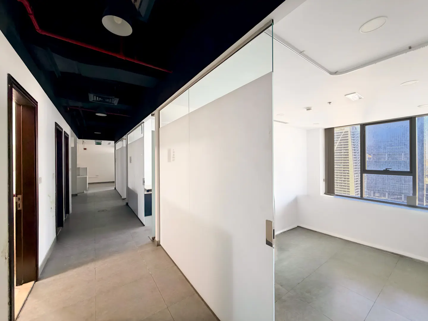 Empty office space with a dark ceiling, white walls, and a gray floor. A window shows a city view.
