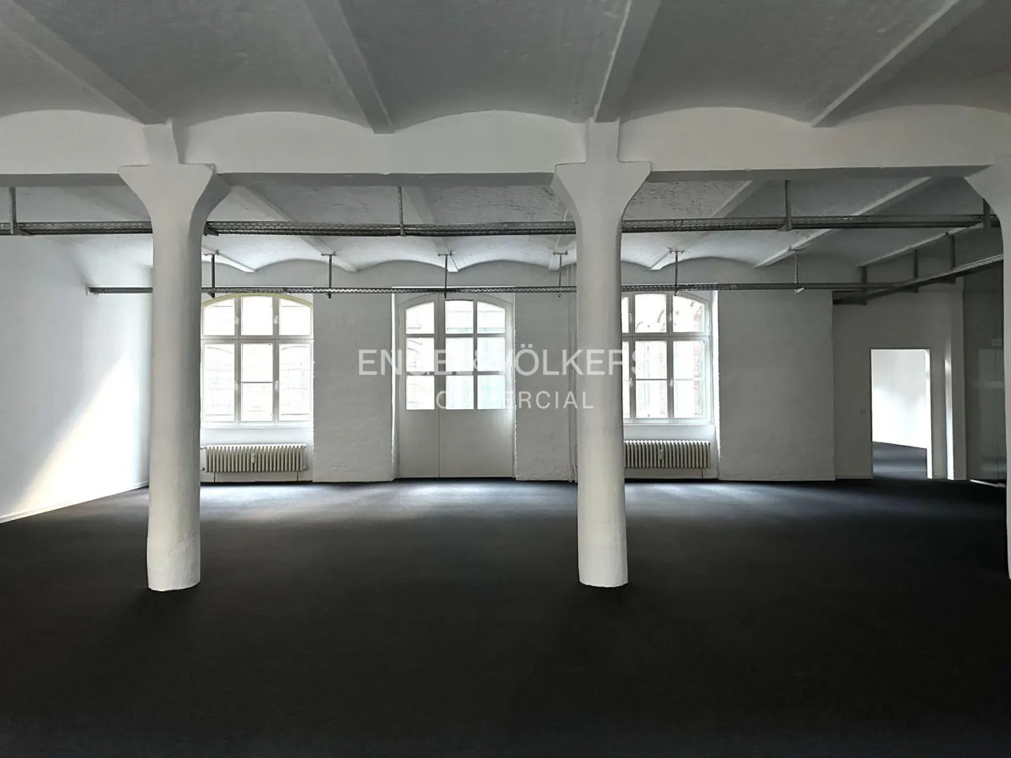 Empty commercial space with white walls, black floor, and large windows. White pillars support the ceiling.