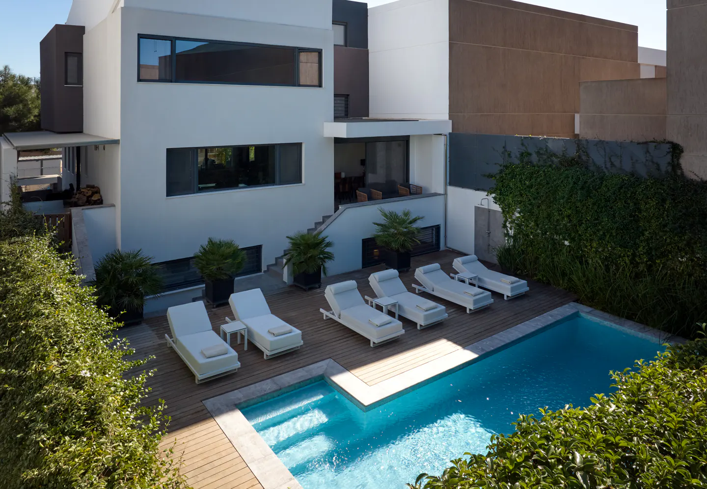 Modern white house with a blue swimming pool, wooden deck, and white lounge chairs. Green bushes surround the pool.