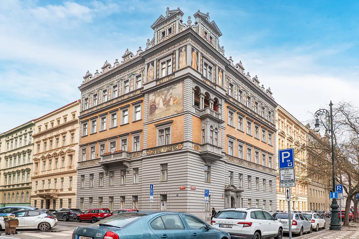 Ornate building with a mural on the corner, gray stone base, orange brick upper floors, and parked cars in front.