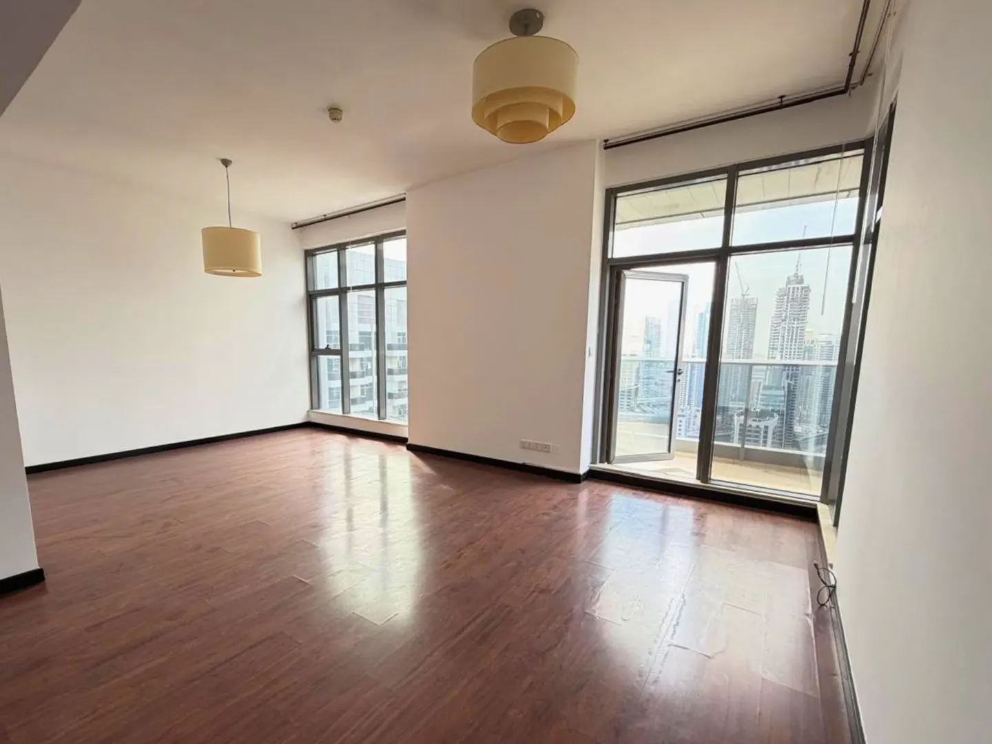 A bright, empty room with hardwood floors, white walls, and large windows with a city view. Two pendant lights hang from the ceiling.