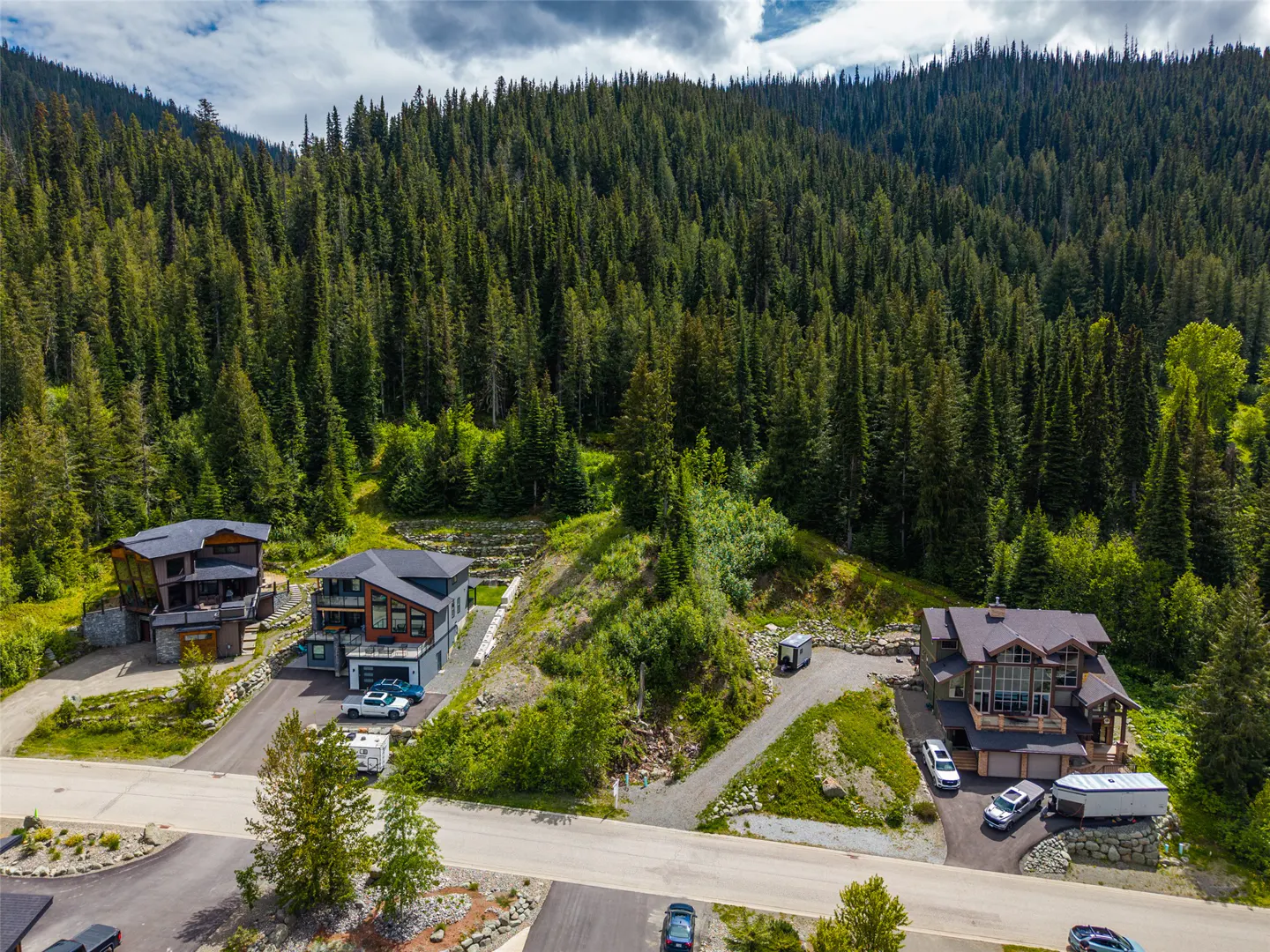 Aerial view of three modern mountain homes nestled in a dense evergreen forest with paved driveways and parked vehicles.