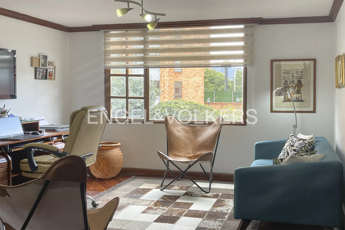 A bright home office with a desk, tan leather chairs, a blue sofa, and a window overlooking a brick building.