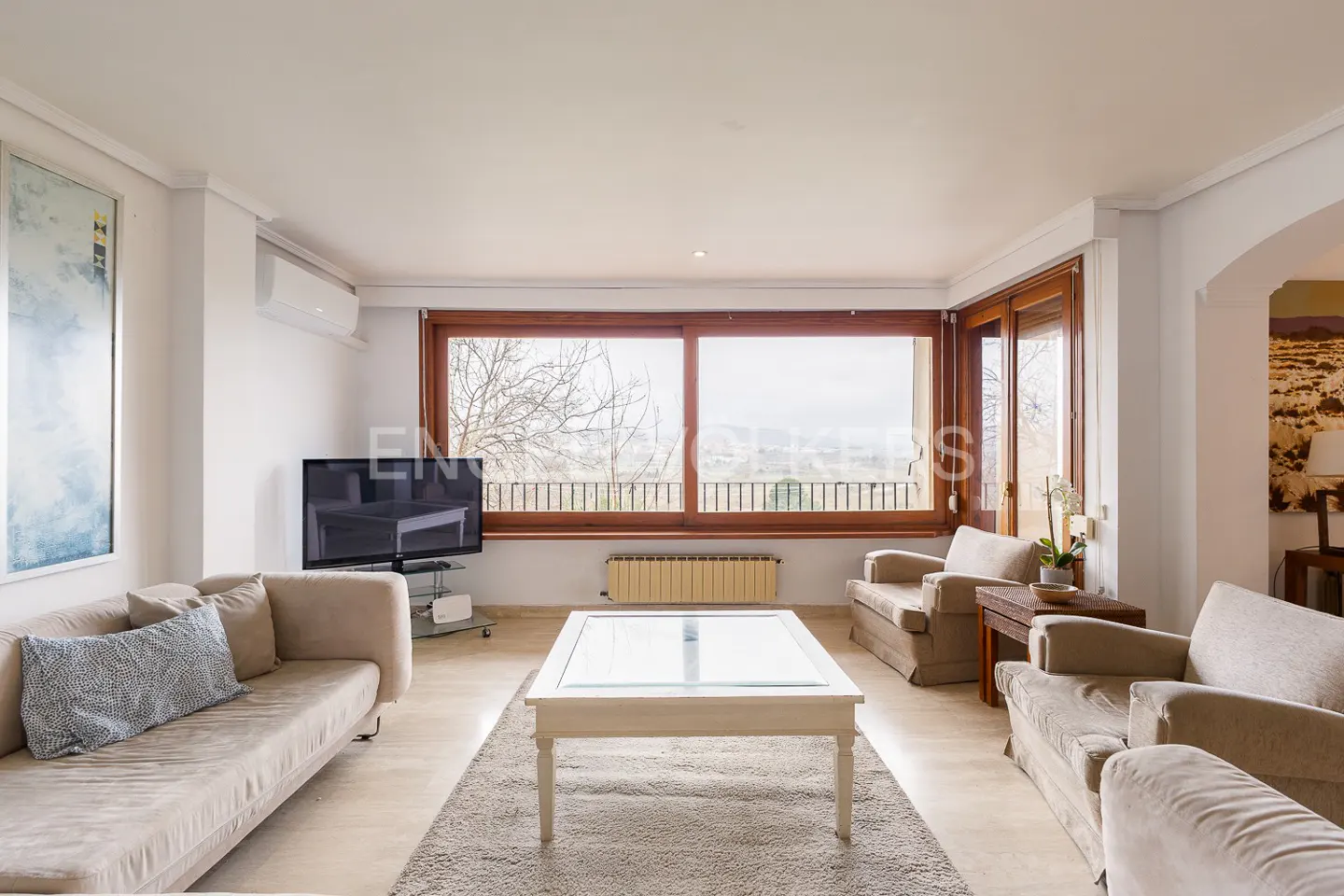 Bright living room with beige sofas, armchairs, and a white coffee table on a gray rug. Large window with a view. TV on a stand.