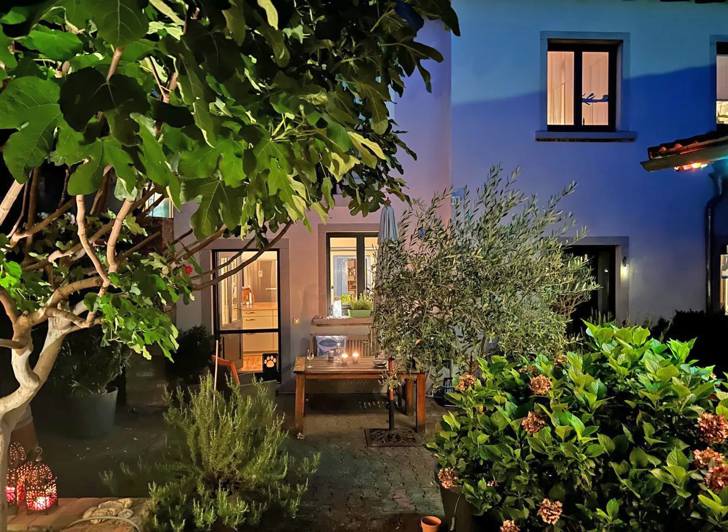 Night view of a home's patio with a wooden table, chairs, plants, and a lantern. The house is white with dark windows.