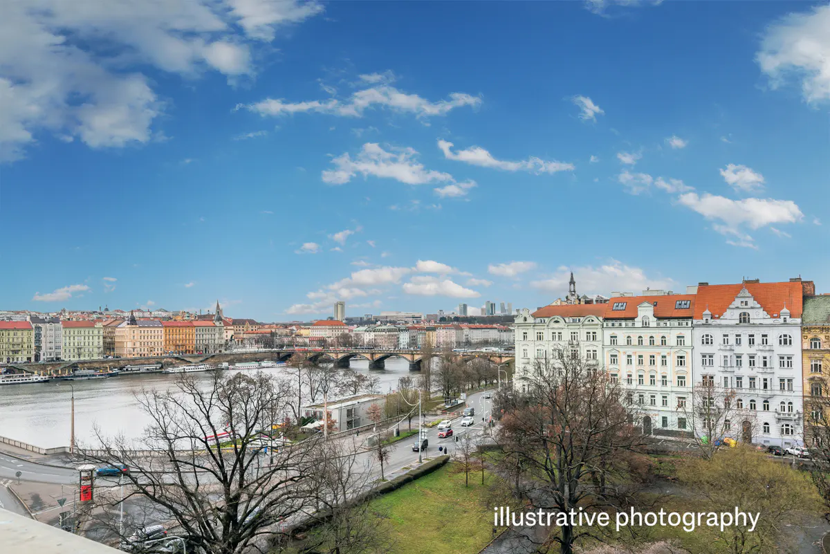 Cityscape view of Prague with the Vltava River, bridges, and colorful buildings under a blue sky with clouds.