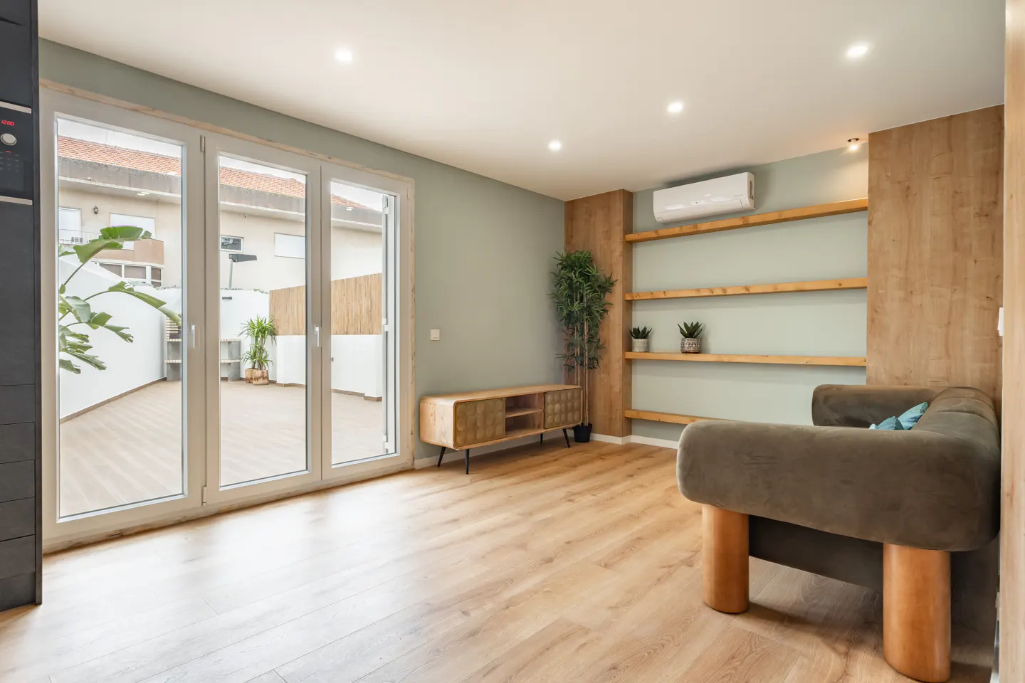 Bright living room with wood floors, green walls, and a large window to a patio. A brown couch and wooden shelves add warmth.