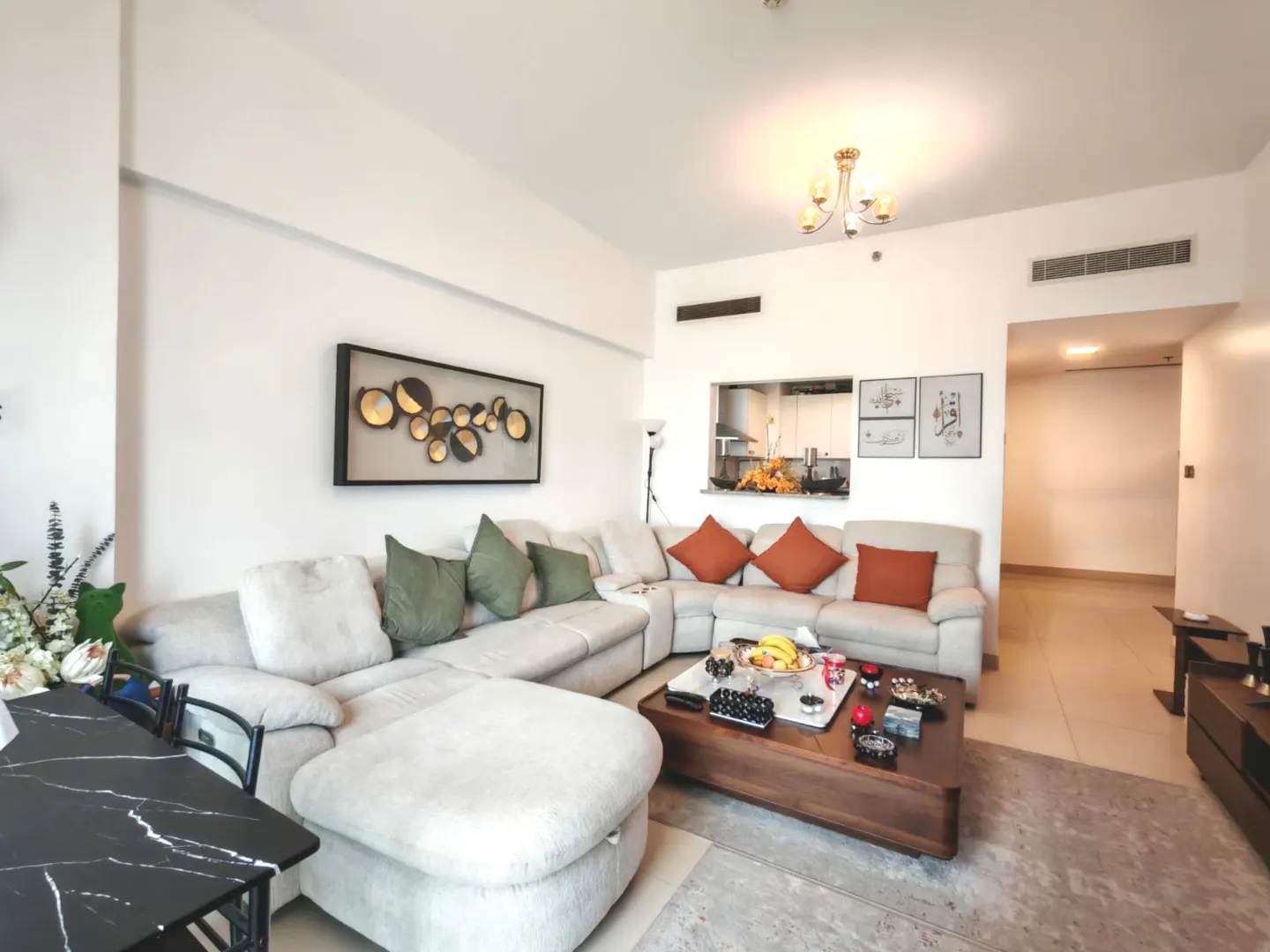 Bright living room with a large, light-colored sectional sofa, a dark wood coffee table, and a black marble dining table. The walls are white, and there is art on the walls.