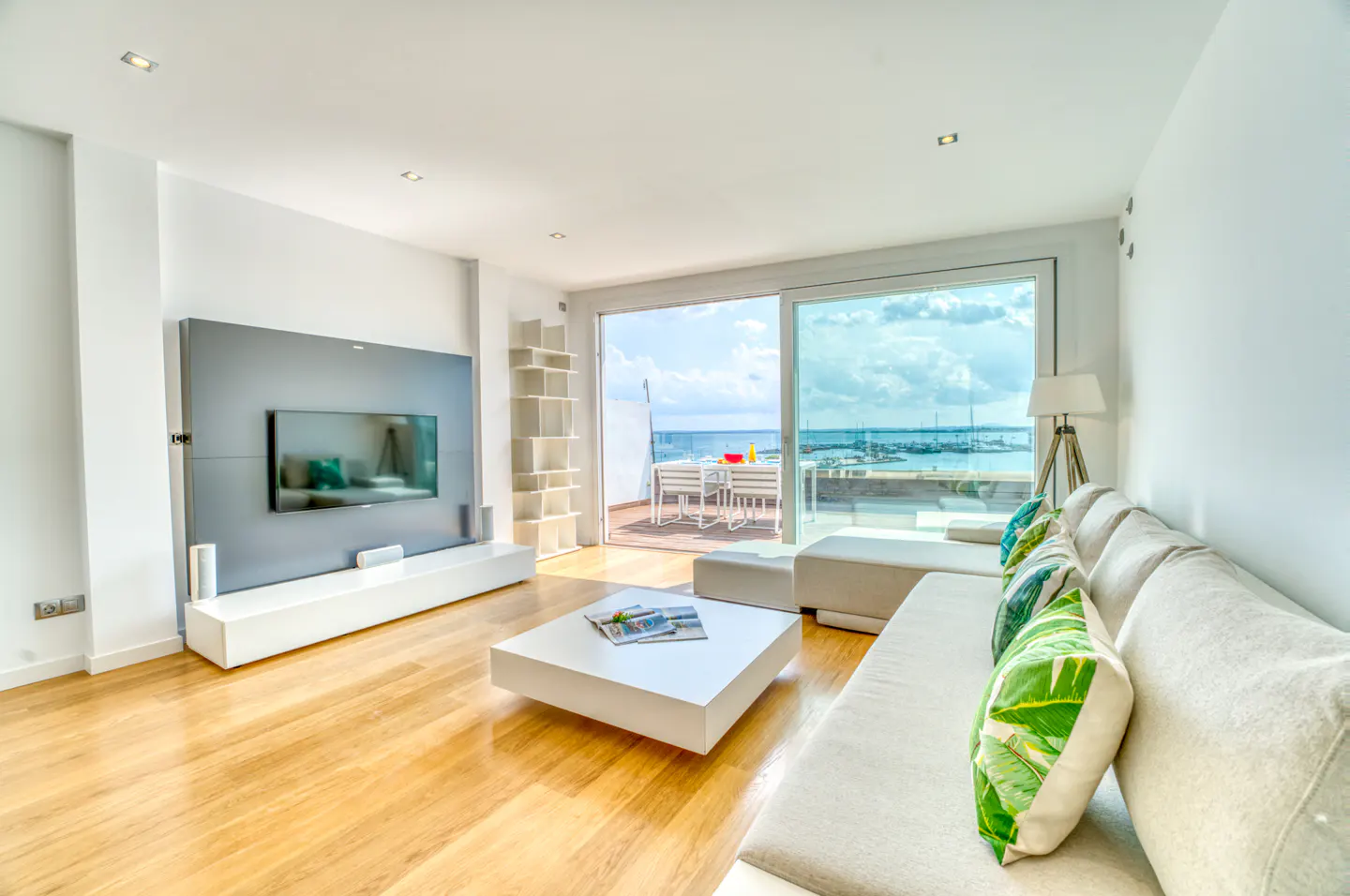 Bright living room with white sofa, square coffee table, and TV. Sliding glass doors open to a deck with ocean views.