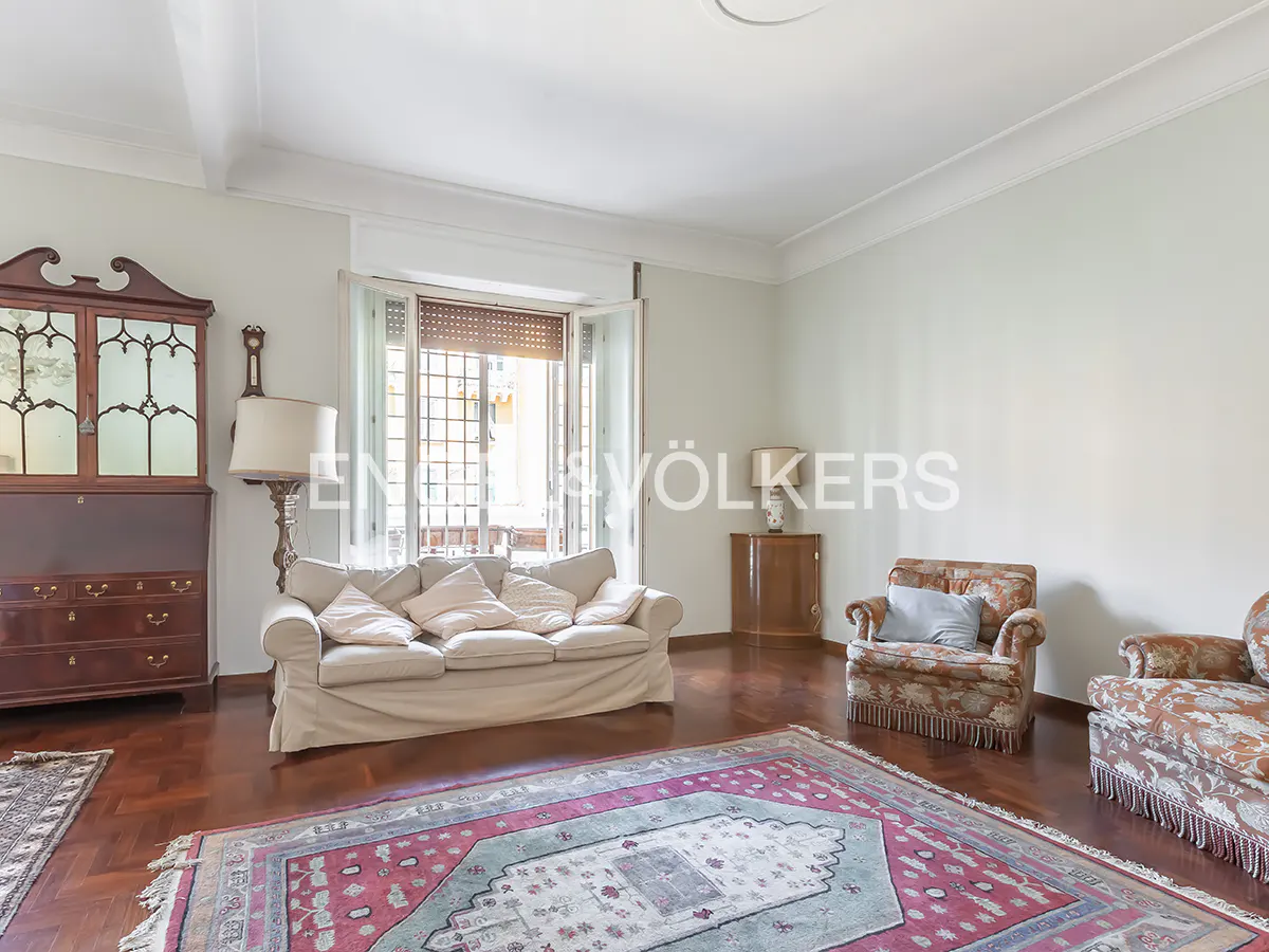 Living room with a beige sofa, two patterned armchairs, a dark wood cabinet, and a large red and teal patterned rug.