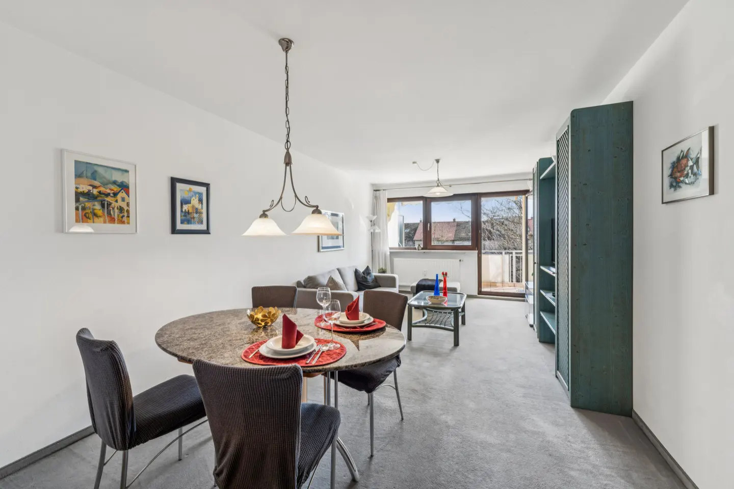 A bright apartment interior with a round dining table set for two, a gray sofa, and a balcony.