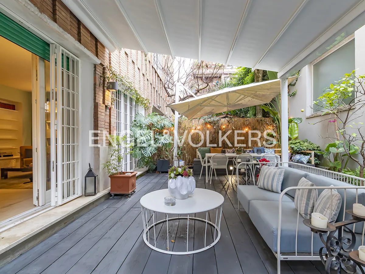 Outdoor patio with gray wood flooring, a light blue sofa, and a white metal coffee table. Lush greenery and a large umbrella provide shade.