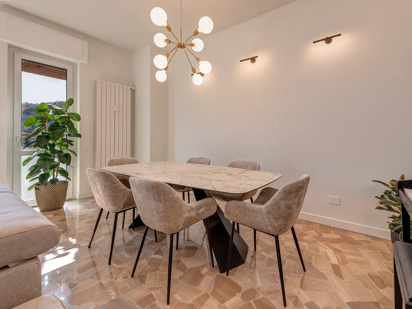 A bright dining room with a marble table, six upholstered chairs, and a modern chandelier. A large plant sits near a window.