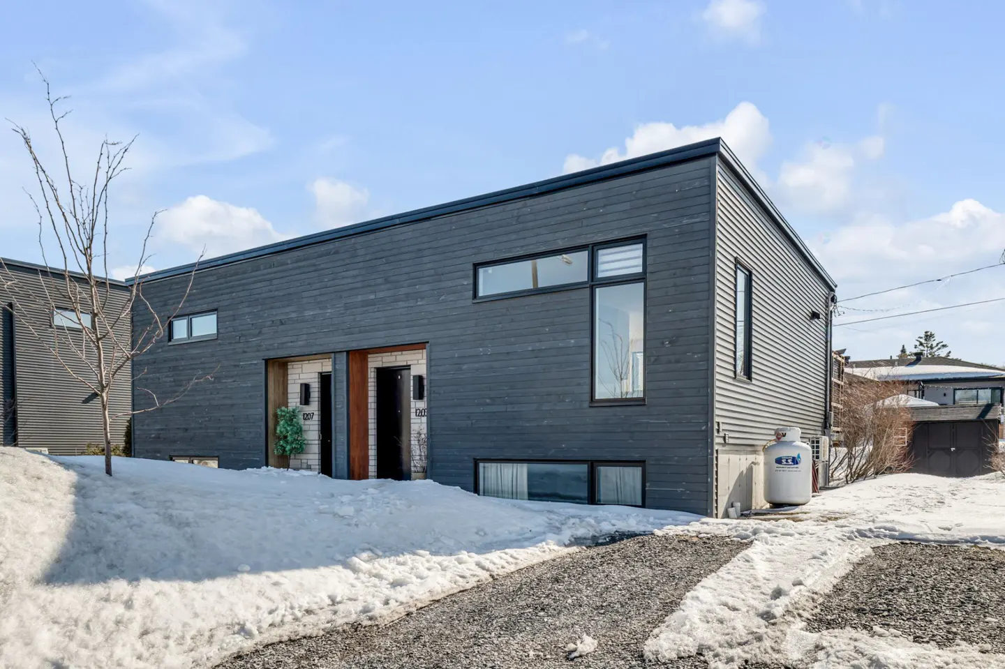 Modern dark gray townhouse with snow-covered yard and gravel driveway under a blue sky.