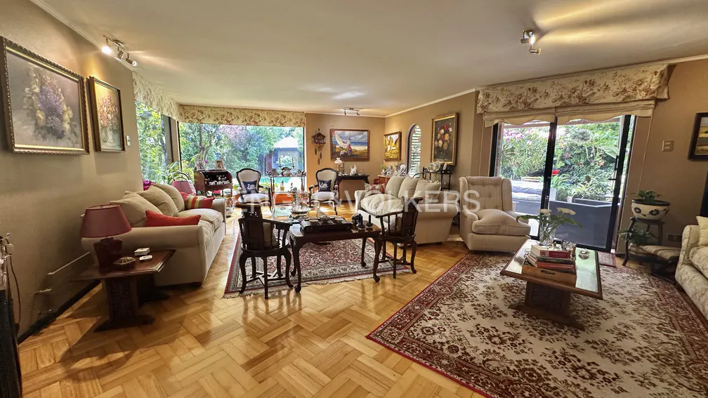 A living room with hardwood floors, sofas, chairs, and rugs. Large windows overlook a garden.