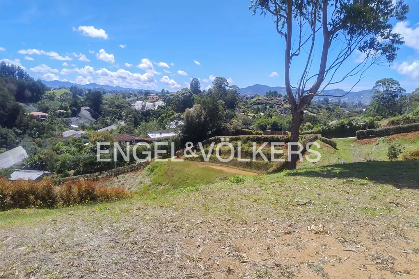 Scenic view of a grassy lot with trees, hills, and houses in the background under a blue sky with clouds. Engel & Volkers logo overlay.