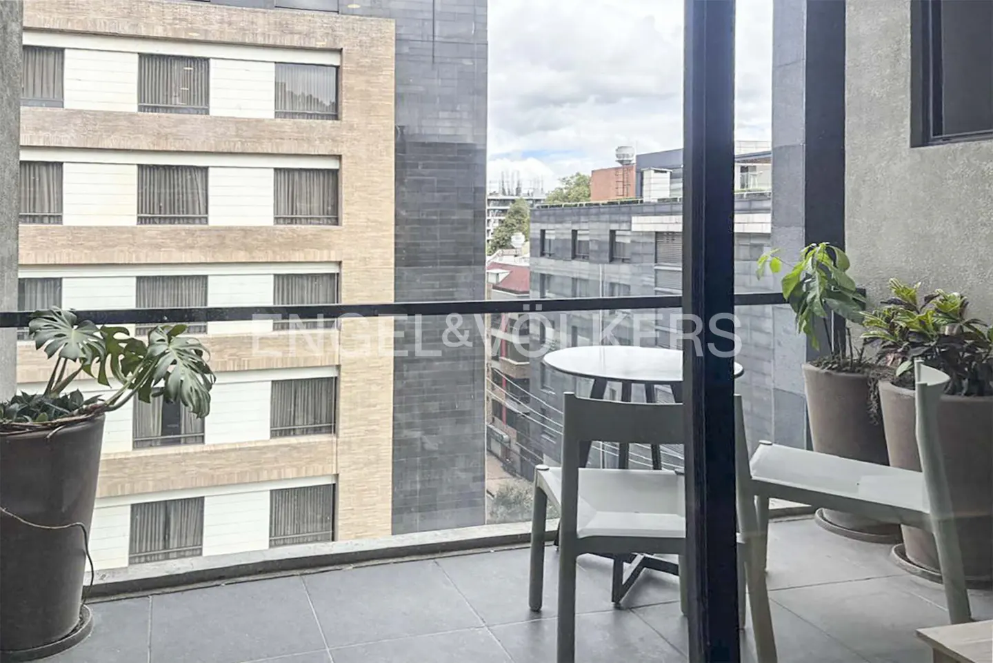 Balcony view with table, chairs, and plants. City buildings visible in the background. Neutral color palette.
