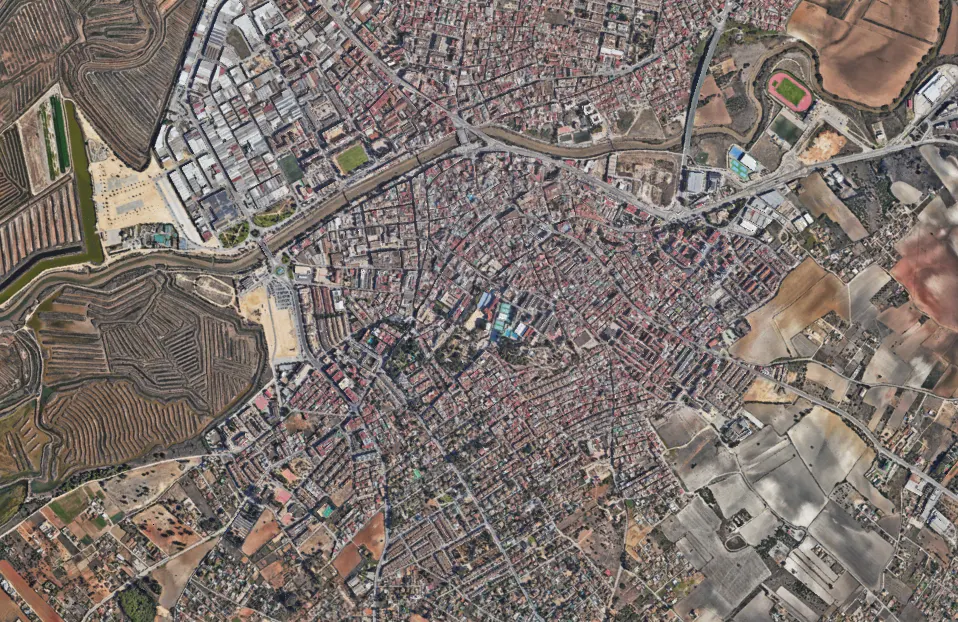Aerial view of a dense city with red-roofed buildings, surrounded by fields and salt marshes. A stadium is visible on the outskirts.