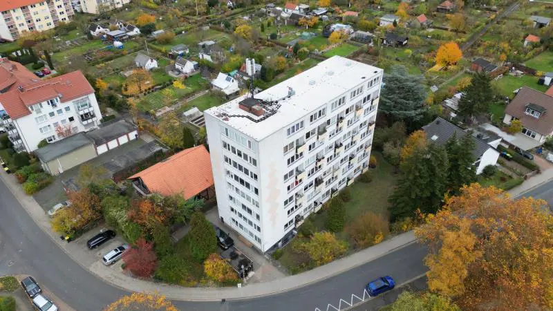 Aerial view of a tall, white apartment building surrounded by houses, trees, and a road in an urban area.