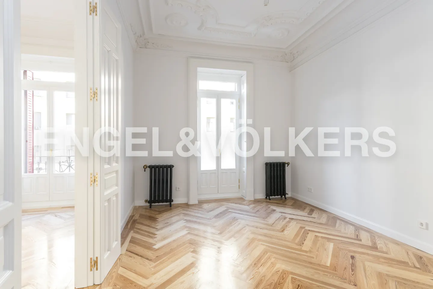 Bright, empty room with herringbone wood floors, white walls, and ornate ceiling. Black radiators flank a window. Doorway to another room is visible.