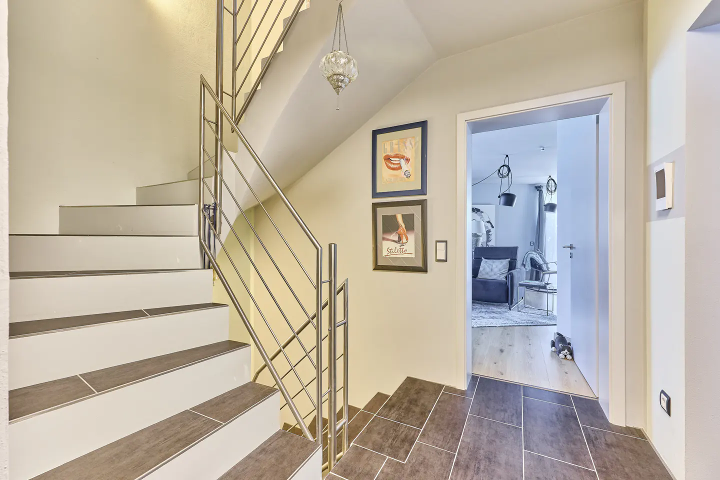 Interior view of a home's staircase with white steps, brown treads, and a stainless steel railing. Artwork adorns the wall, and an open doorway reveals a living room.