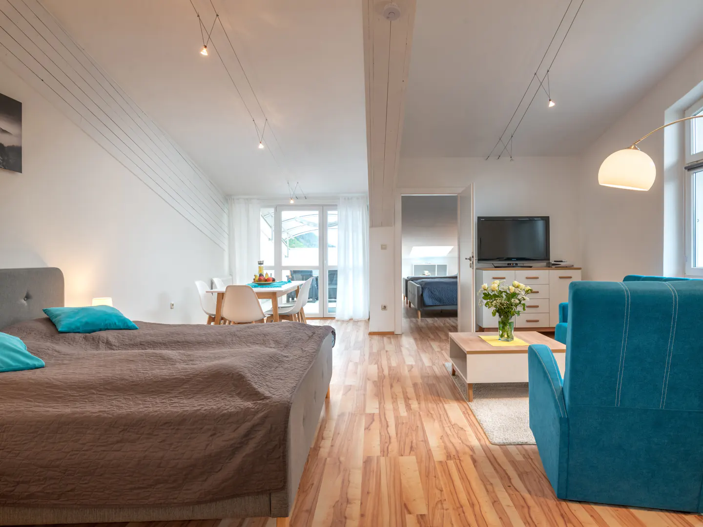 Bright, open-concept apartment with a bed, dining table, blue chairs, and a TV. The walls are white, and the floors are wood.