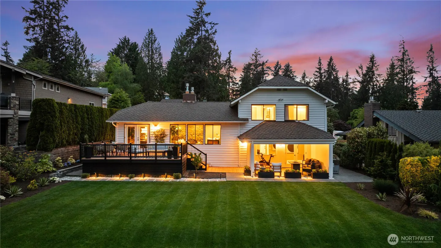 Back of a white two-story house with a deck and green lawn at dusk. Trees surround the property.