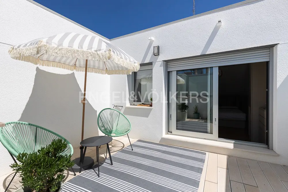 Outdoor patio with white walls, blue sky, striped umbrella, green chairs, and a gray striped rug. Sliding glass doors lead inside.