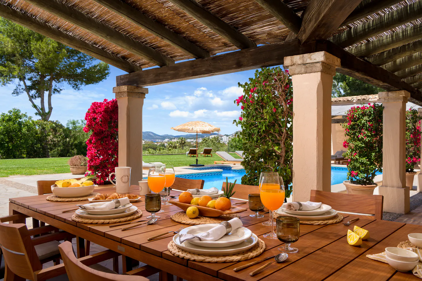 Outdoor dining table set for breakfast on a patio overlooking a swimming pool, lush garden, and distant hills.