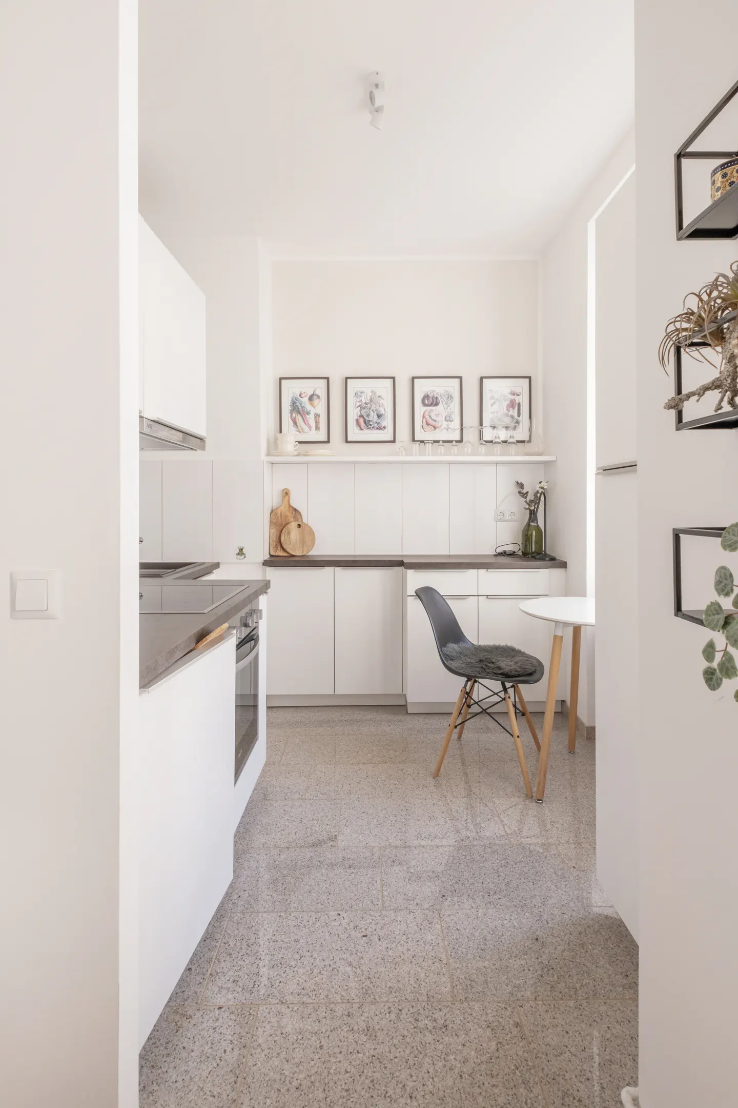 Bright kitchen with white cabinets, dark countertops, and gray speckled floor. A gray chair sits near a small white table. Wall art and shelves add detail.