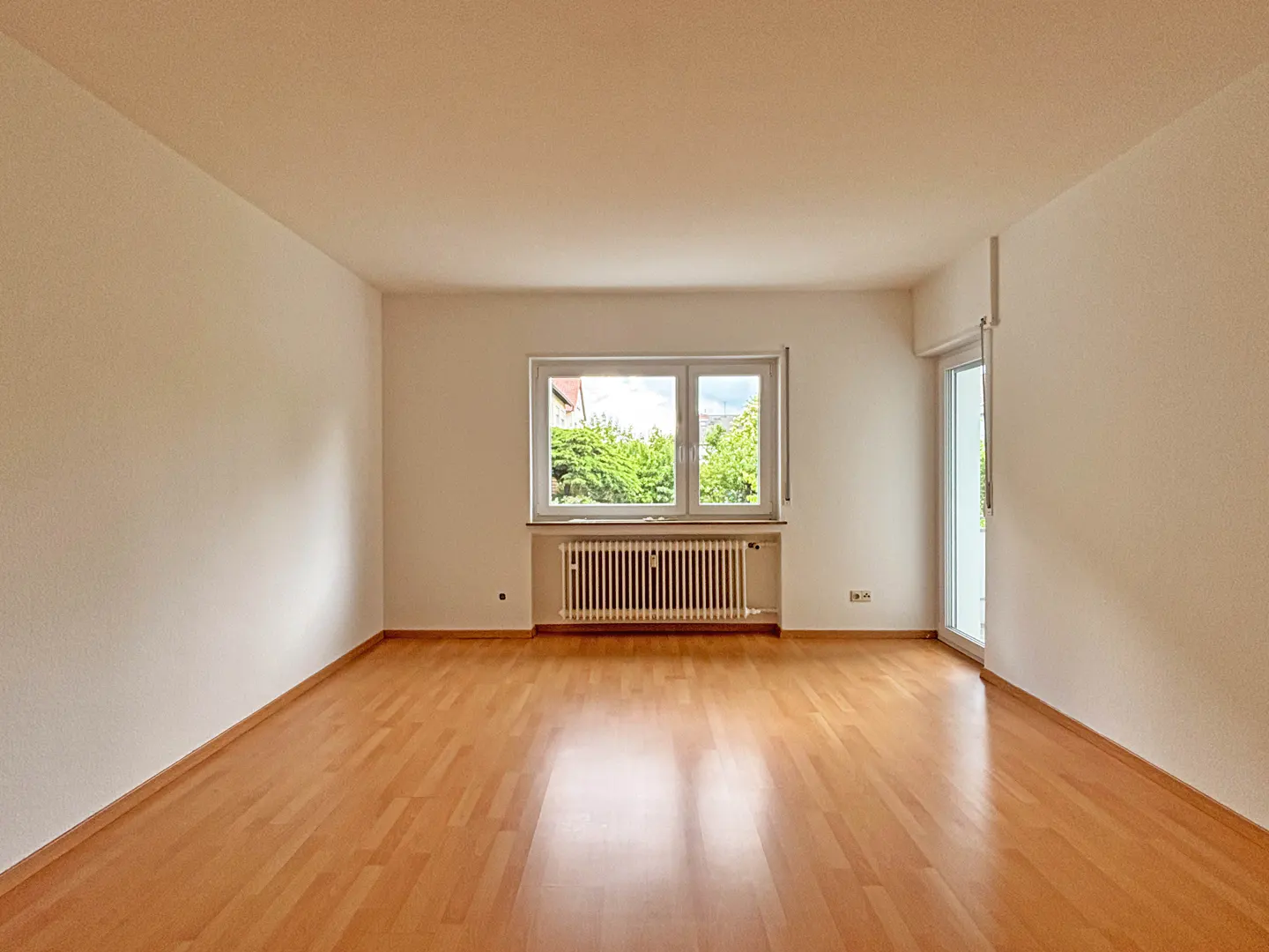 Empty room with light wood floors, white walls, and a window overlooking green trees. A white radiator sits below the window.