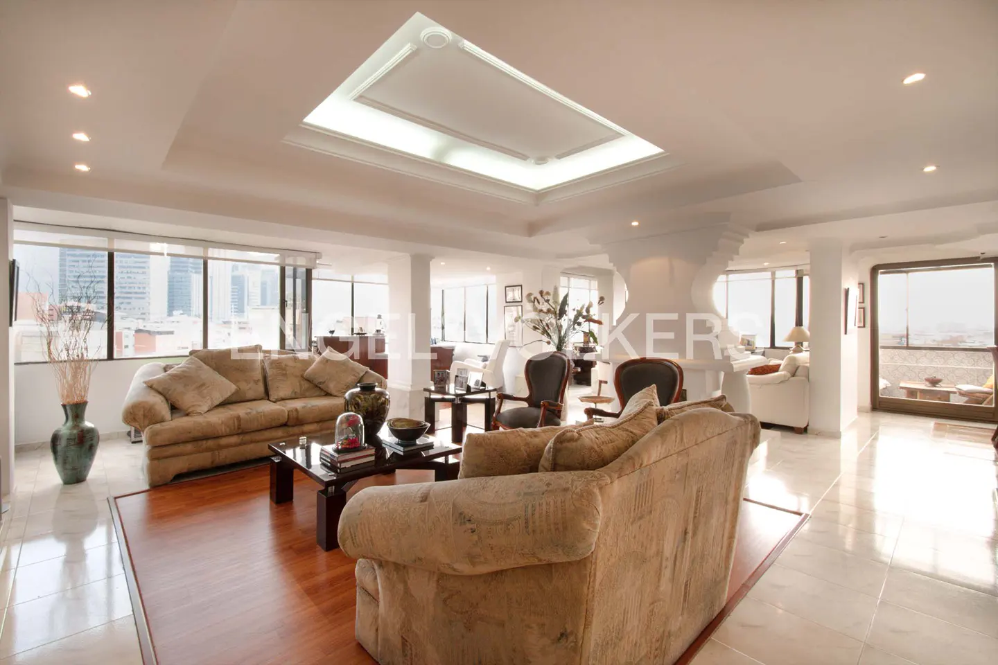 Bright, open living room with beige sofas, dark wood tables, and a city view through large windows.