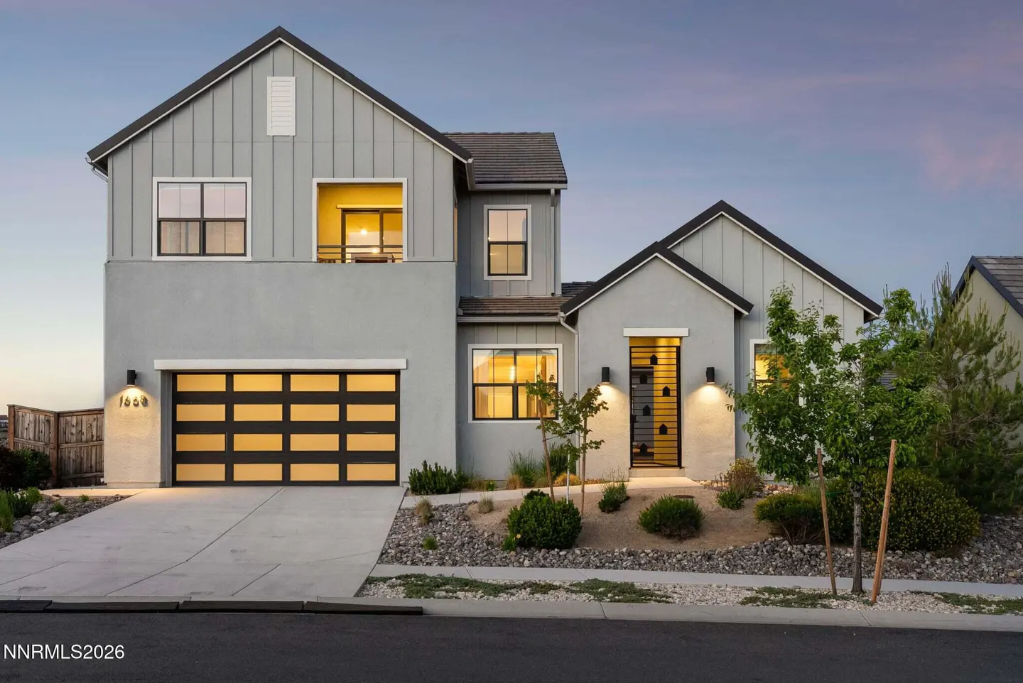 Modern two-story gray home with a garage, lit windows, and desert landscaping at dusk.