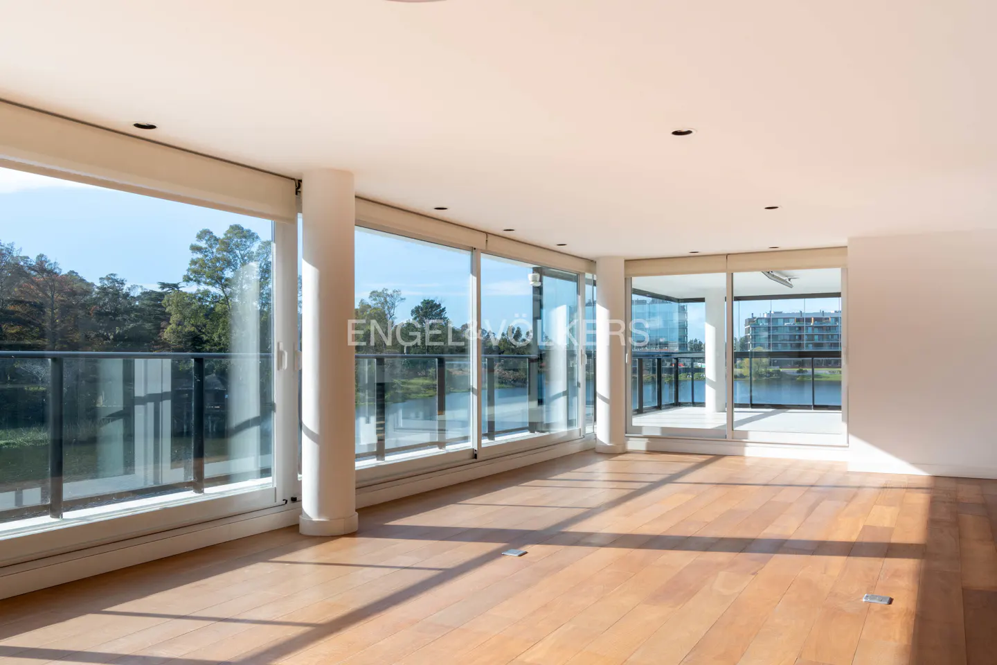 Bright, empty room with wood floors and floor-to-ceiling windows overlooking a lake and trees. Sunlight streams in.